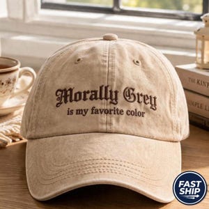 Może przedstawiać: Beżowa czapka z daszkiem o wykończeniu z postarzanego materiału. Z przodu znajduje się ciemnobrązowy haft z napisem Morally Grey is my favorite color wykonanym ozdobną czcionką szeryfową. Czapka posiada usztywnianą koronę i zakrzywiony daszek.