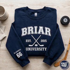 Op de afbeelding: Marineblauwe sweatshirt met witte tekst "BRIAR UNIVERSITY EST. 2015" en gekruiste hockeysticks. De mouw heeft de naam "DI LAURENTIS" en het nummer "66". Een hockeypuck en -stick zijn zichtbaar.