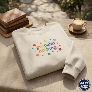 May include: A cream-coloured sweatshirt embroidered with the phrase "probably nothing" in rainbow hues, encircled by floral and star motifs. The sweatshirt rests on a wooden table, with books and a coffee cup visible in the background.
