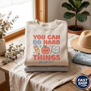 May include: A beige crewneck sweatshirt featuring a graphic with the text "YOU CAN DO HARD THINGS" in red and blue, accompanied by illustrations of a pencil, a smiling apple, and an open book, with the smaller text "you got this!" below.