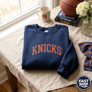 Può includere: Una felpa girocollo blu navy con la scritta KNICKS ricamata in un carattere arcuato con lettere arancioni e un contorno blu. Il capo presenta maniche lunghe e colletto, polsini e orlo a coste, caratterizzato da un design sportivo.