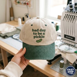 May include: A beige and green baseball cap with the text "bad day to be a pickle" and a pickle graphic. The cap has a curved brim and adjustable strap. The hat is a casual accessory.