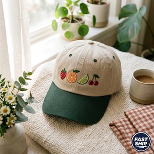 May include: A beige baseball cap with a dark green brim. The cap features embroidered fruit designs, including a strawberry, orange slice, lime wedge, and cherries. The cap is sitting on a cream-colored blanket.