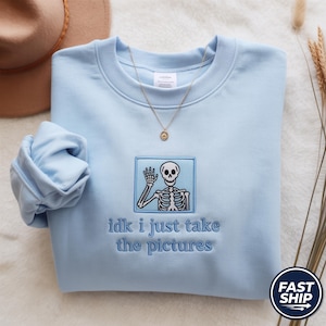 May include: A light blue crewneck sweatshirt featuring an embroidered square patch with a skeleton waving its hand. Below the patch, the text reads, "idk i just take the pictures" in dark blue thread. A gold-toned necklace rests on the collar.