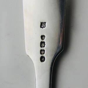 May include: Close-up of the back of a silver spoon with hallmarks. The hallmarks include a crown, the letters "MP", a shield, and a lion.