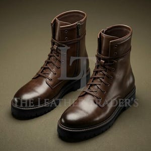 Può includere: Un paio di stivali in pelle marrone con suole nere. Gli stivali hanno lacci, cerniere e un cinturino alla caviglia. Il testo "THE LEATHER TRADER'S" è visibile sul lato degli stivali.