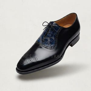 May include: A black leather oxford shoe featuring a polished finish, a blue crocodile-embossed leather panel on the lace-up section, thin black laces, and decorative brogue perforations on the rounded toe cap. The text THE LEATHER TRAD is visible.