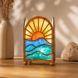 May include: An arched table lamp with a wooden frame featuring a stained glass-style panel. The panel displays a sunrise over ocean waves in shades of orange, yellow, light blue, and teal. The lamp stands on four small wooden ball feet.
