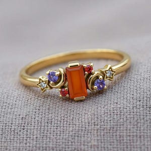 May include: A gold ring featuring a central rectangular orange stone, flanked by two crescent moon shapes, two small purple round stones, two tiny red round stones, and two small white star-shaped stones arranged along the thin gold band.