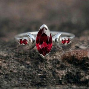 May include: A silver ring with a large, marquise-cut, deep red gemstone as the focal point. The ring features crescent moon designs on either side of the central stone, each with a smaller red stone. The ring is set against a textured, gray background.