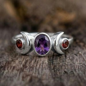May include: A silver ring featuring a central oval-cut purple amethyst gemstone flanked by two crescent moon shapes, each containing a small round-cut red garnet stone. The metal band has a polished finish and a symmetrical design.