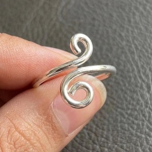 Può includere: Un anello bypass color argento caratterizzato da due motivi a spirale aperti sulla parte anteriore. La fascia è liscia e lucida, con le due estremità che si curvano in distinti vortici circolari rivolti in direzioni opposte.