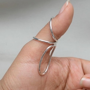 May include: A silver-colored metal splint ring featuring a smooth, continuous wire design that wraps around the thumb joint. The structure consists of two circular bands connected by a vertical loop, providing support to the finger joint.