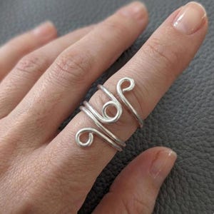 May include: A silver-colored wire ring with a unique, swirling design. The ring features multiple loops and curves, creating an artistic and eye-catching accessory. The ring is worn on a finger, showcasing its intricate details and craftsmanship.
