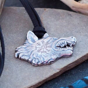 May include: A silver wolf head pendant with a compass design on the forehead, hanging on a black leather cord. The pendant is resting on a gray stone with a blue and black feather in the background.
