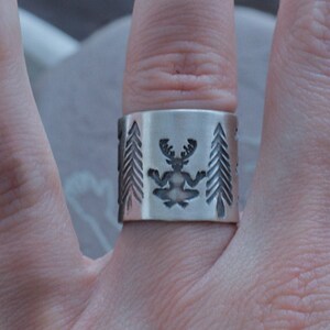May include: A silver ring with a design of a deer with antlers and pine trees on either side.