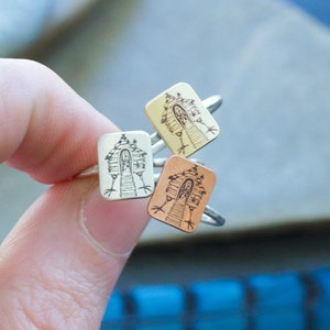 May include: Three silver rings with a square design featuring a hand-drawn illustration of a house. The rings are stacked on top of each other, with the top ring being silver, the middle ring being gold, and the bottom ring being copper.