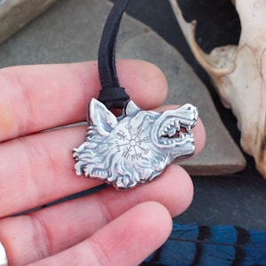 May include: Silver wolf head pendant necklace with a black cord. The pendant features a detailed wolf head with a circular design inside. The pendant is held in a hand with a ring on the finger.