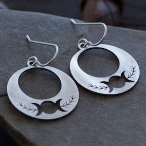 May include: A pair of silver earrings with a triple moon design and leafy branches. The earrings are hanging from a silver hook.