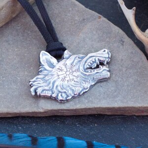 May include: Silver wolf head pendant with a compass design on the forehead, hanging from a black cord on a gray stone surface. A blue and black feather and a white animal skull are also in the image.