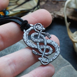 May include: A silver-colored pendant necklace featuring a double snake design. The pendant is on a black cord. The background includes a butterfly, a skull, and a piece of wood.