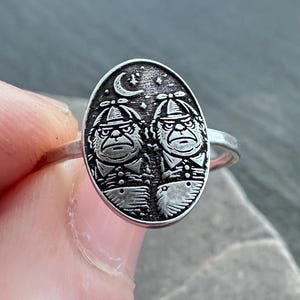 May include: Silver ring featuring an oval-shaped face with two figures under a crescent moon and stars. The ring has a detailed design with a vintage or antique aesthetic. The ring is held by a person's fingers.