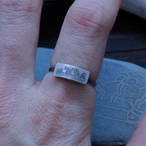 May include: A silver ring with a rectangular face featuring a detailed landscape design. The ring is worn on a finger, showcasing the intricate craftsmanship. The ring's band is thin, and the design is in a minimalist style.