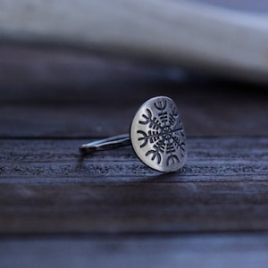 May include: A silver ring with a circular design featuring a snowflake-like pattern. The ring is lying on a wooden surface.