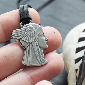 May include: Silver pendant necklace featuring a profile of a woman with a winged headdress. The pendant is on a black leather cord.