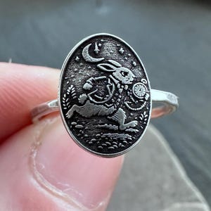 May include: Silver ring with an oval face. The face has a detailed engraving of a rabbit wearing a waistcoat and holding a pocket watch, set against a starry night sky. The ring's band has a textured design.