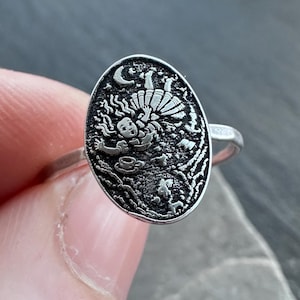 May include: Silver ring with an oval-shaped face featuring a detailed, engraved design. The design includes a figure, a crescent moon, and other intricate elements. The ring is held between fingers, showcasing its polished finish and craftsmanship.