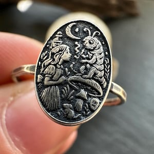 May include: Silver ring with an oval pendant featuring a detailed engraving. The design depicts a scene with a girl, a caterpillar, a crescent moon, and other elements. The ring is held by a person.