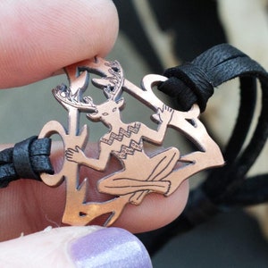 May include: A copper-colored bracelet with a detailed design of a horned figure. The bracelet is made of leather cord and features a pendant with a stylized human figure with antlers.