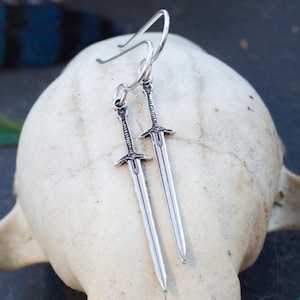 May include: A pair of silver sword earrings hanging from a white skull. The swords are detailed and have a slightly aged look.