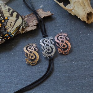 May include: Three silver, copper, and gold snake knot pendants on a black leather cord. The pendants are in the shape of a snake knot, a traditional Celtic symbol.