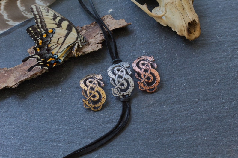 May include: Three silver, copper, and gold snake knot pendants on a black leather cord. The pendants are in the shape of a snake knot, a traditional Celtic symbol.