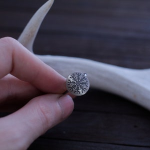 May include: A silver ring with a Vegvisir symbol engraved on the face. The ring is being held by a hand against a wooden background.