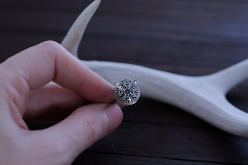 May include: A silver ring with a Vegvisir symbol engraved on the face. The ring is being held by a hand against a wooden background.