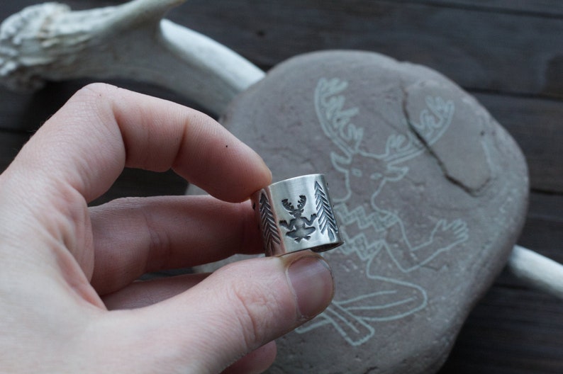 May include: A silver ring with a detailed design of a deer with antlers and pine trees. The ring is being held by a hand.