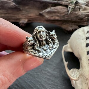 K&ouml;nnte beinhalten: Ein silberner Ring mit dem Gesicht einer Frau mit langen Haaren, flankiert von zwei Katzen. Der Ring weist aufwendige Details und eine Vintage-&Auml;sthetik auf. Der Ring wird von einer Hand gehalten.