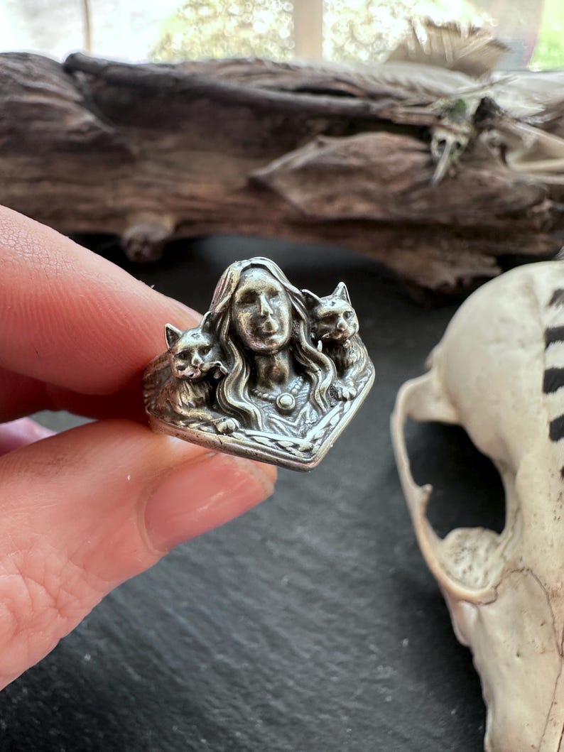 K&ouml;nnte beinhalten: Ein silberner Ring mit dem Gesicht einer Frau mit langen Haaren, flankiert von zwei Katzen. Der Ring weist aufwendige Details und eine Vintage-&Auml;sthetik auf. Der Ring wird von einer Hand gehalten.