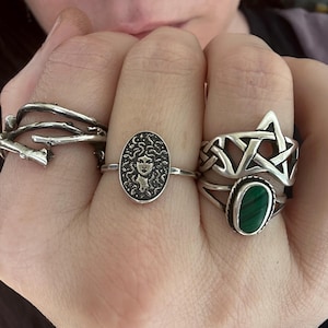 May include: A hand displays four silver rings. One ring features a branch design, another has an oval face, and a third has a green stone. The rings are detailed and have an antique or gothic aesthetic.