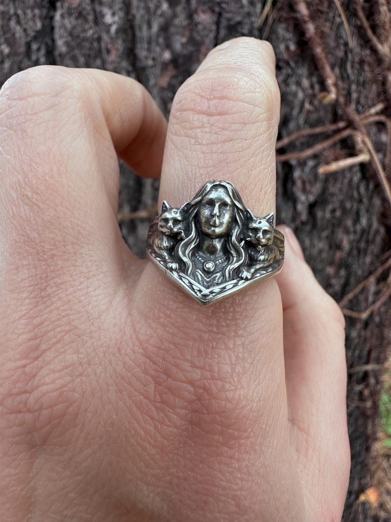 K&ouml;nnte beinhalten: Ein silberner Ring mit einer zentralen Frauenfigur und zwei Katzenfiguren. Der Ring hat ein aufwendiges, gotisches Design. Die Fassung ist dreieckig. Der Ring ist aus Silber gefertigt.