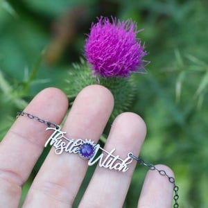 May include: A silver necklace with a purple gemstone and the words "Thistle Witch" in cursive.