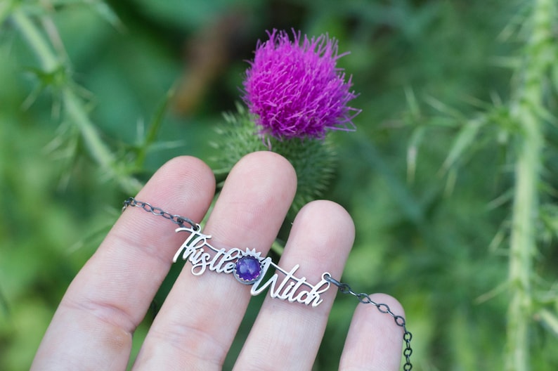 May include: A silver necklace with a purple gemstone and the words "Thistle Witch" in cursive.