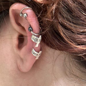 May include: Silver ear cuff with a bird design and the word "NNX" engraved on it. The ear cuff is worn on the upper part of the ear, above a silver hoop earring with a black bead.