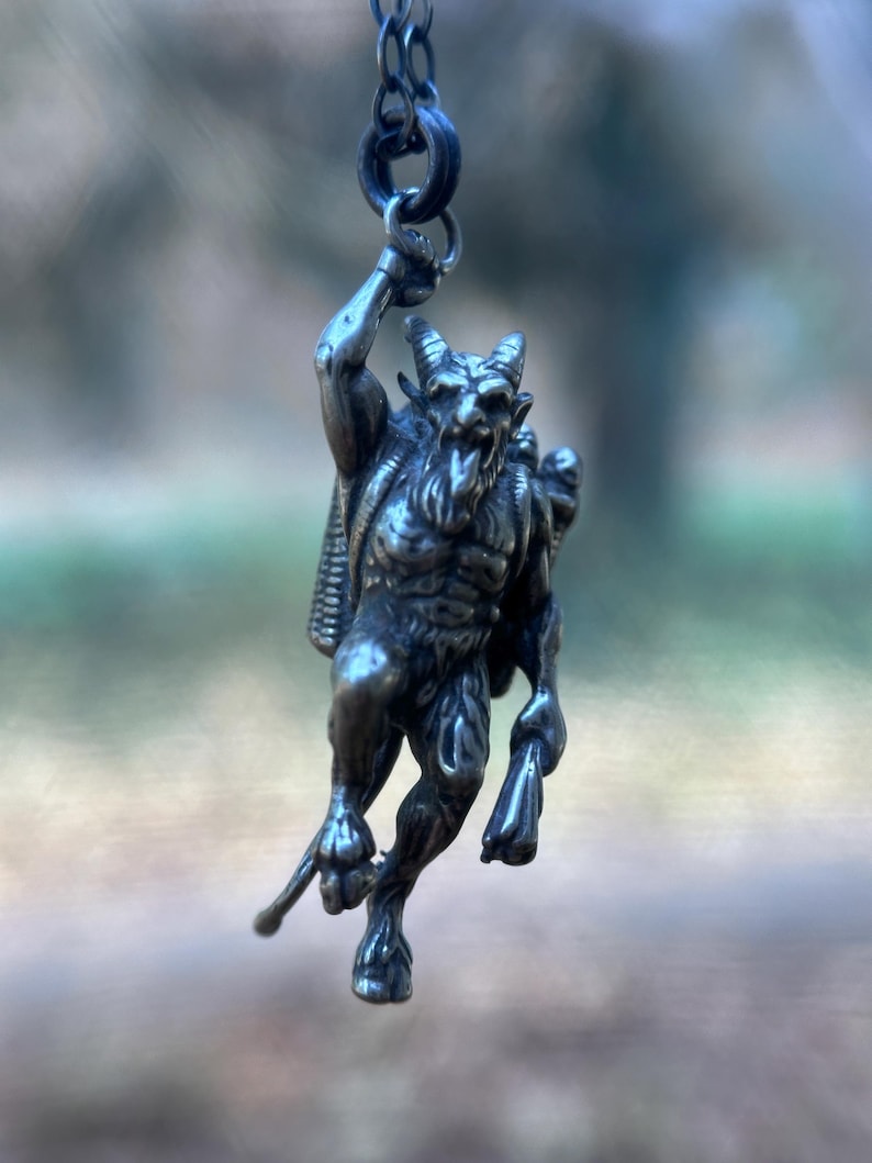 May include: A silver-toned pendant depicting a horned, muscular figure with a beard and a satchel. The pendant is suspended from a chain, showcasing intricate details of the mythical creature. The figure is holding a staff.