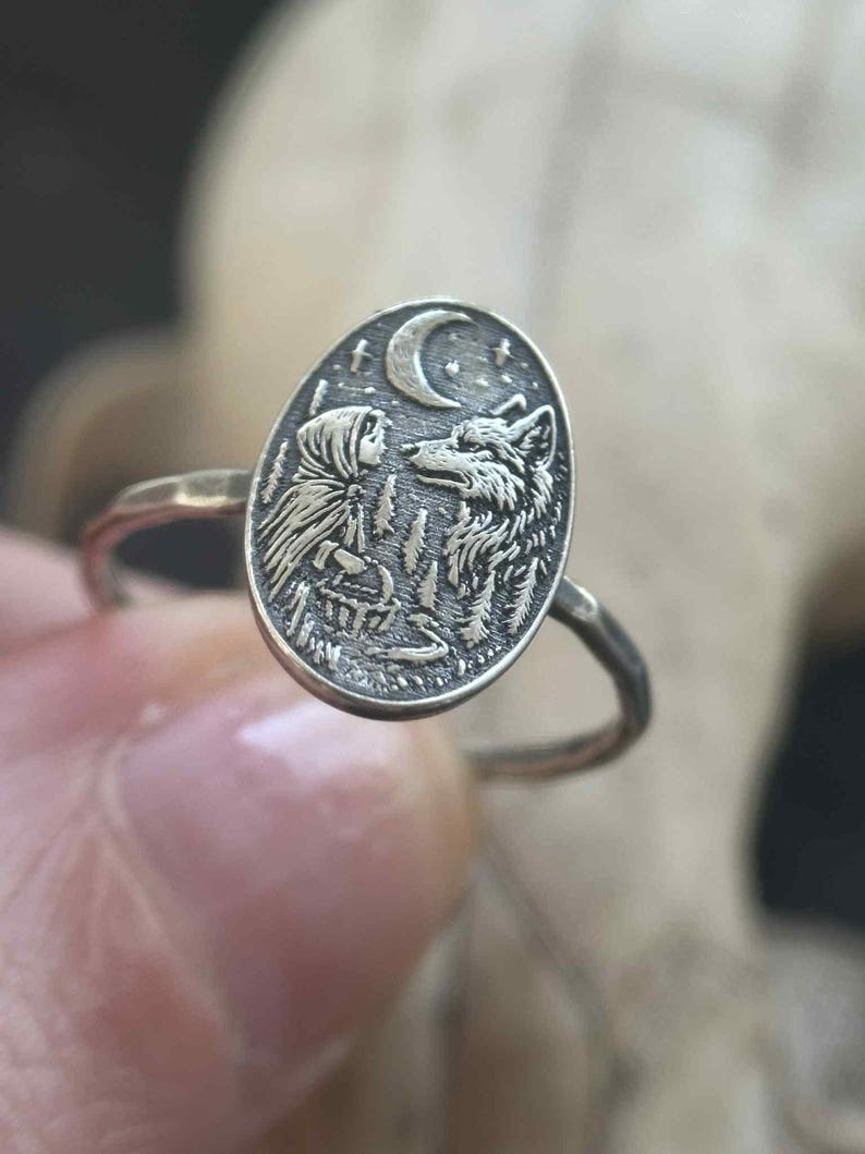 May include: Silver-toned ring featuring an oval pendant with a detailed relief of a person and a wolf under a crescent moon. The ring's design includes forest elements, creating a nature-inspired theme. The ring is held by a person's hand.