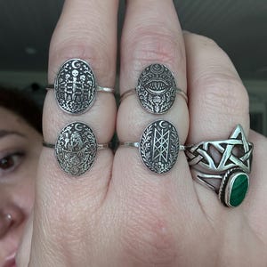 May include: A collection of silver rings with various designs. One ring features a five-pointed star with a green stone. Other rings have oval faces with detailed patterns, including skeletons, eyes, and geometric shapes. The rings are displayed on a hand.