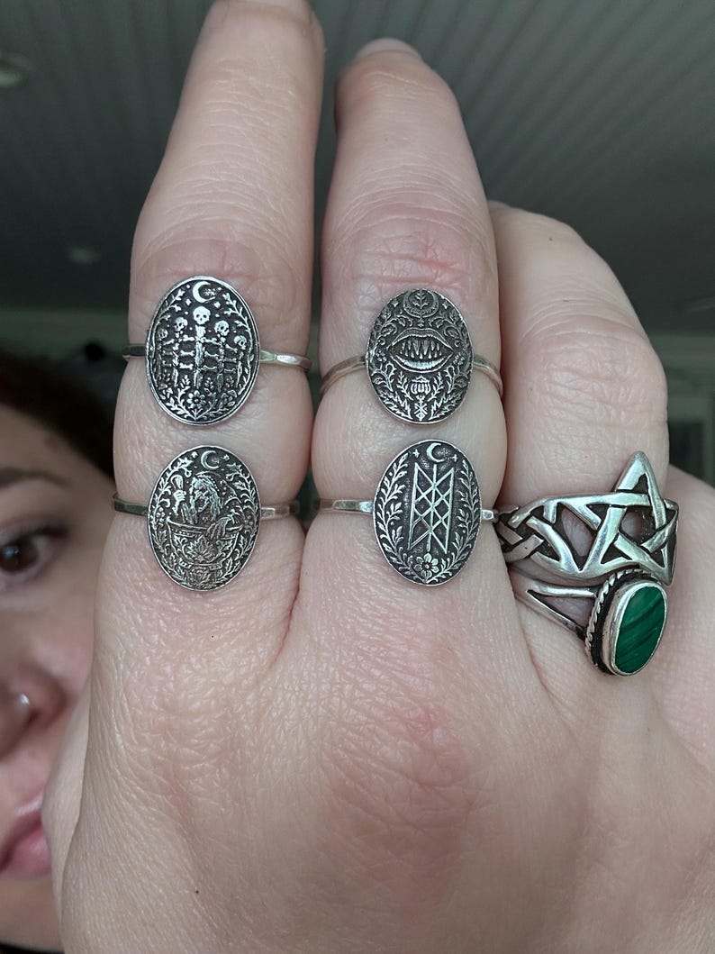 May include: A collection of silver rings with various designs. One ring features a five-pointed star with a green stone. Other rings have oval faces with detailed patterns, including skeletons, eyes, and geometric shapes. The rings are displayed on a hand.